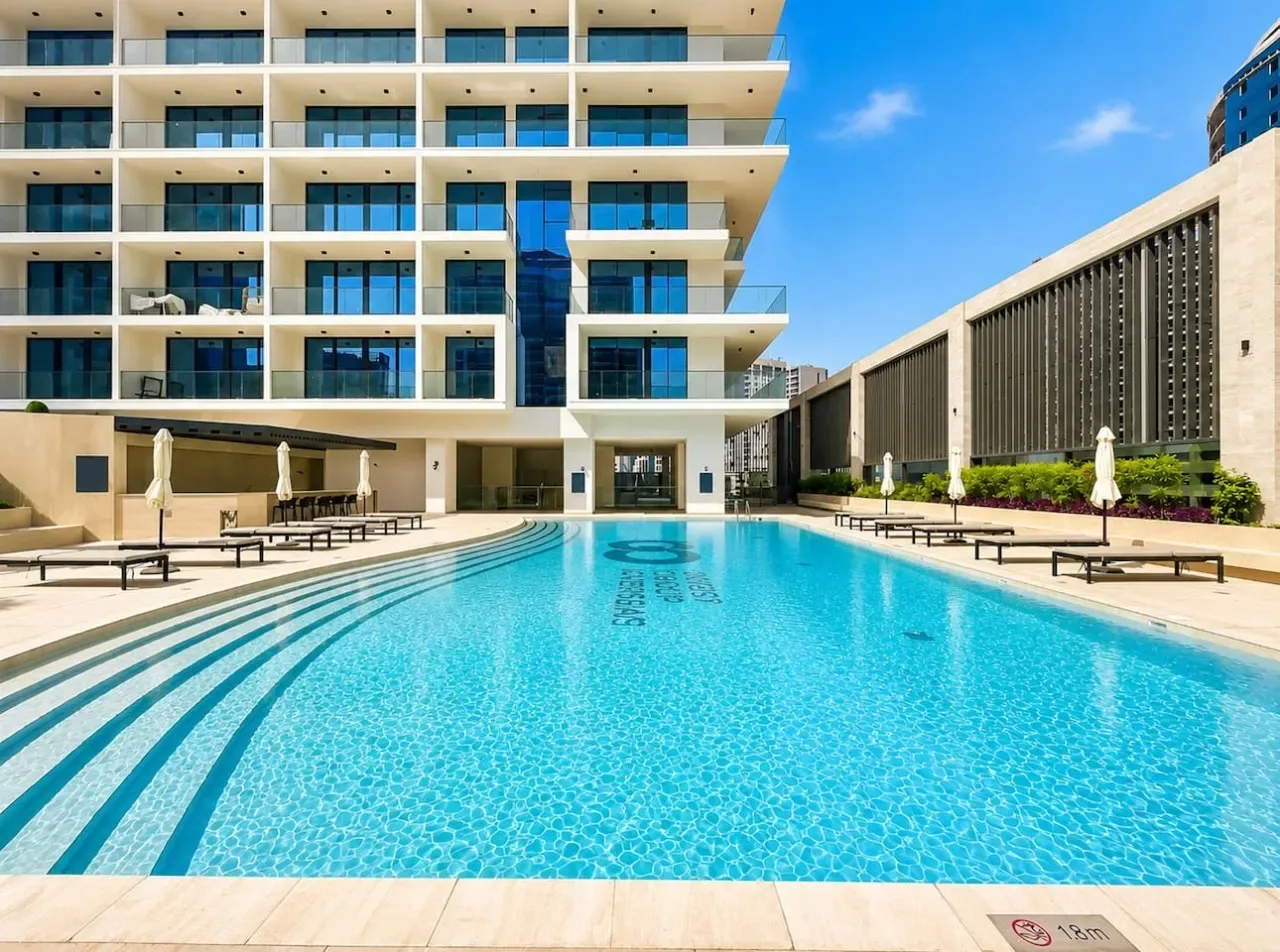 Una piscina está junto a un edificio moderno de varias plantas bajo un cielo azul despejado.