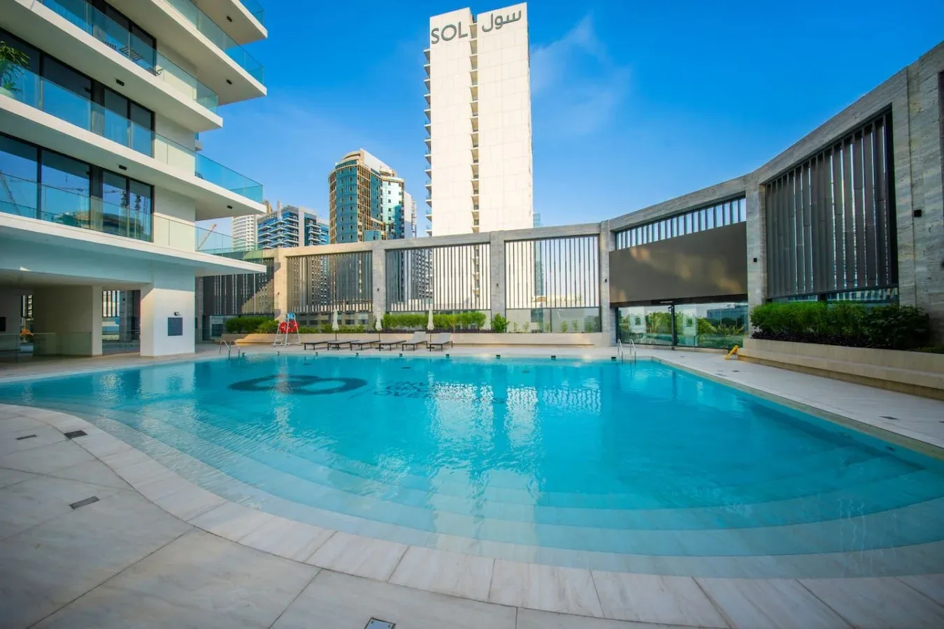 An outdoor swimming pool sits among modern buildings under a clear blue sky.