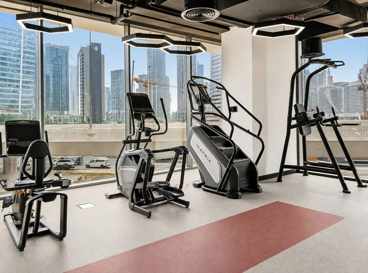 A gym with exercise machines beside large windows overlooking city buildings.