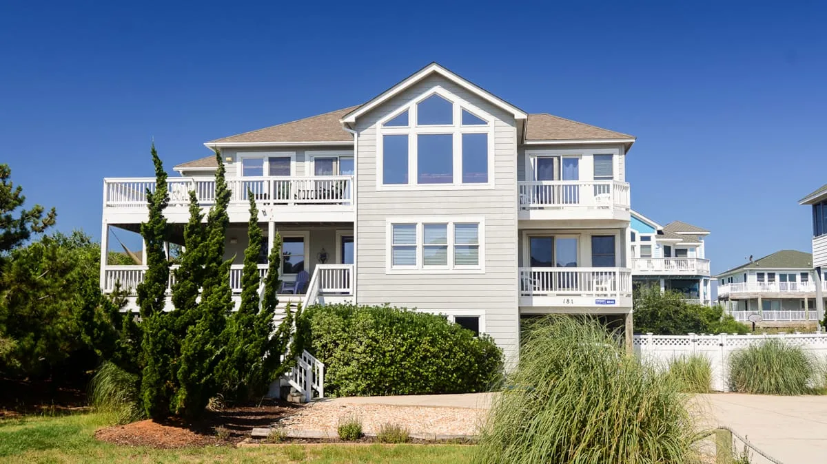 A large light-colored house with balconies stands behind landscaped greenery.