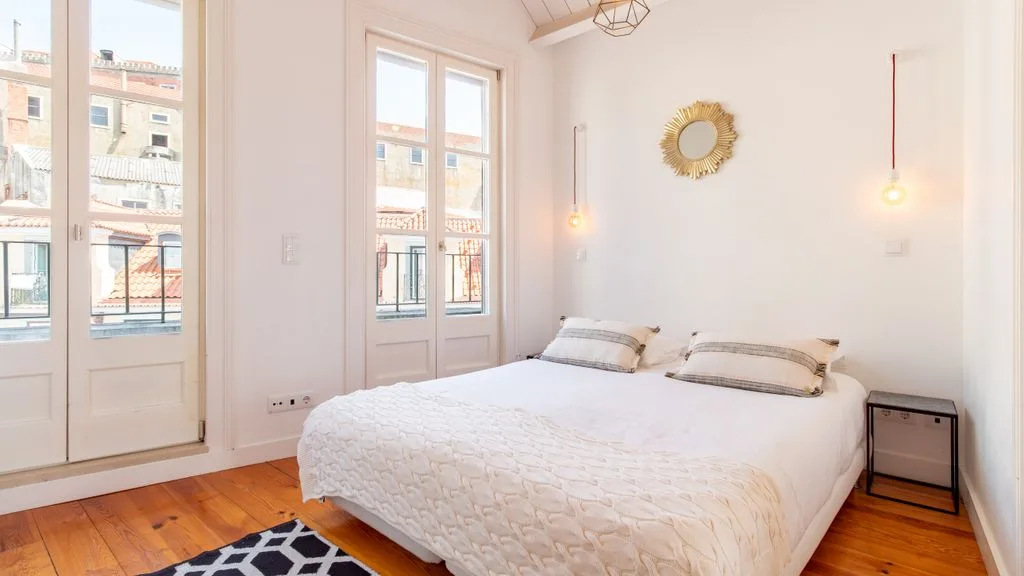 Un dormitorio luminoso con una cama junto a ventanas altas que dan a un balcón.