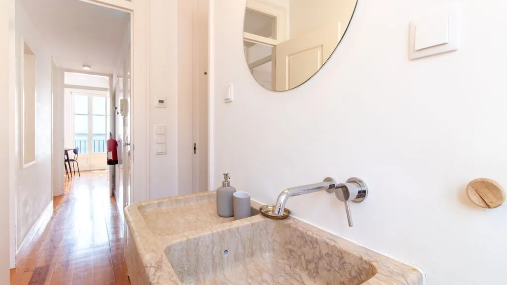 Un baño luminoso con lavabo de piedra, grifo de pared y espejo redondo.