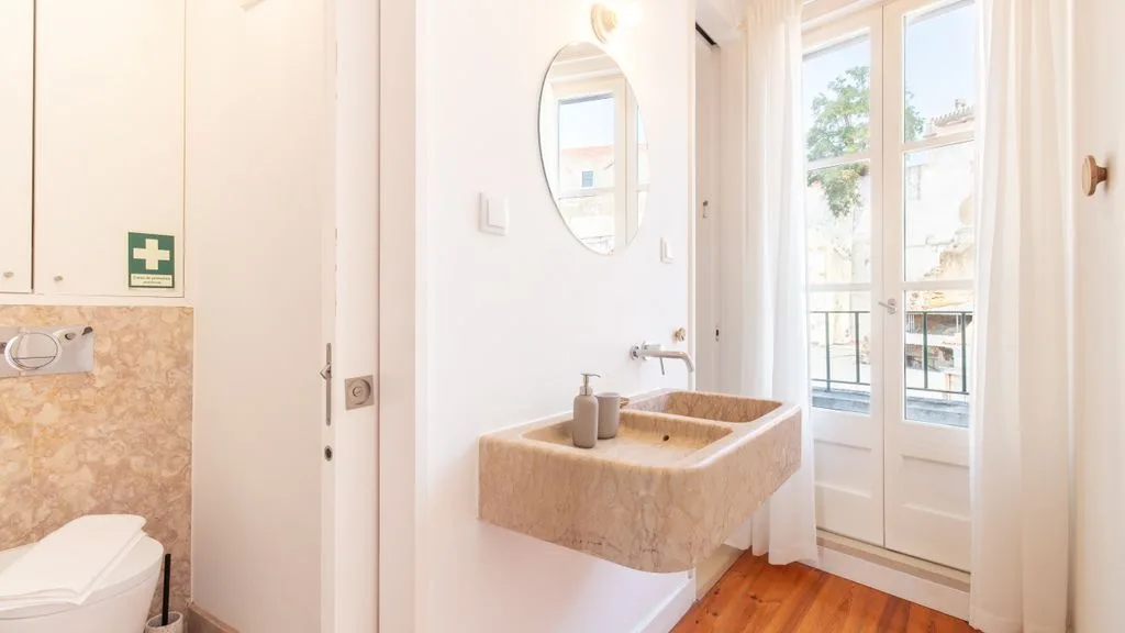 Un baño luminoso con lavabo de piedra y altas puertas de cristal que dan a un balcón.