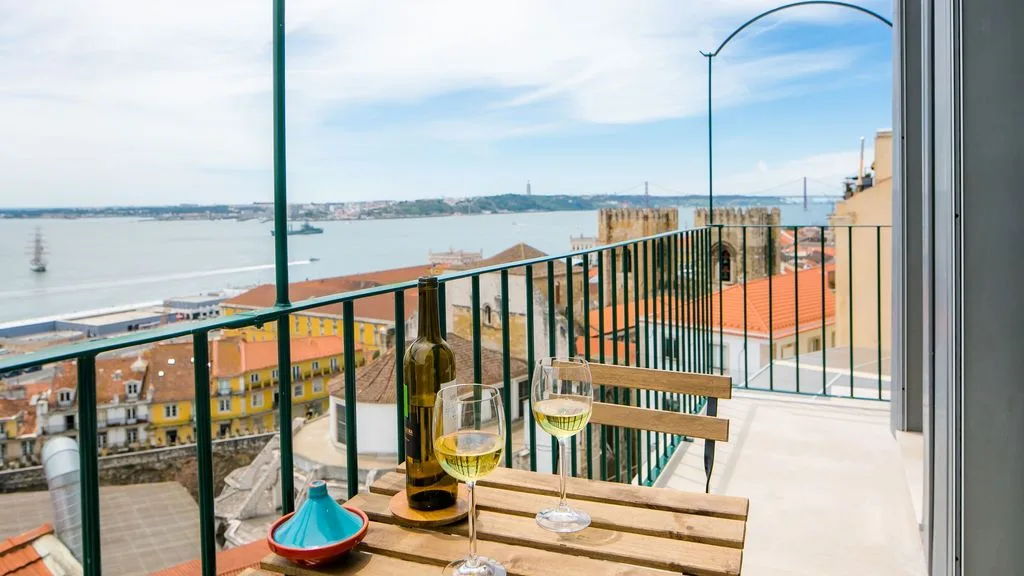 Una mesa de balcón con copas mira hacia un barrio costero y una amplia bahía.