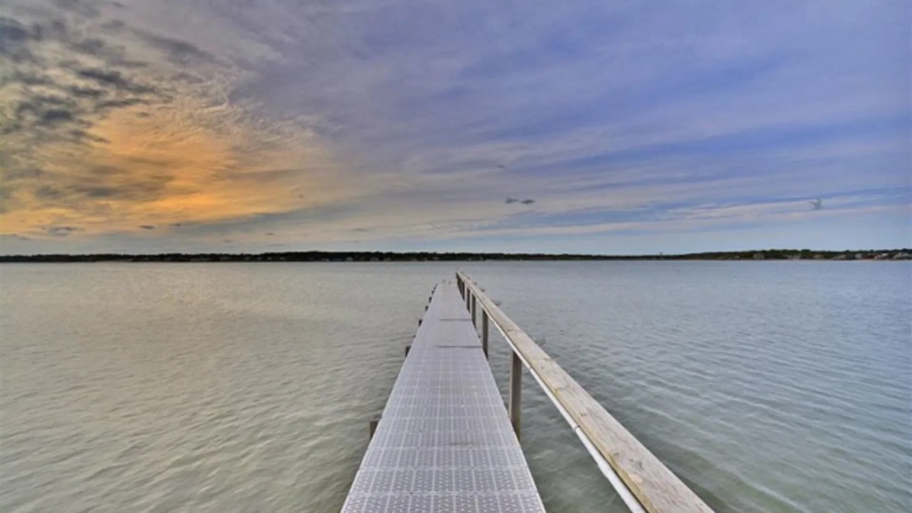 Un muelle estrecho se extiende sobre aguas tranquilas bajo un cielo nublado al atardecer.