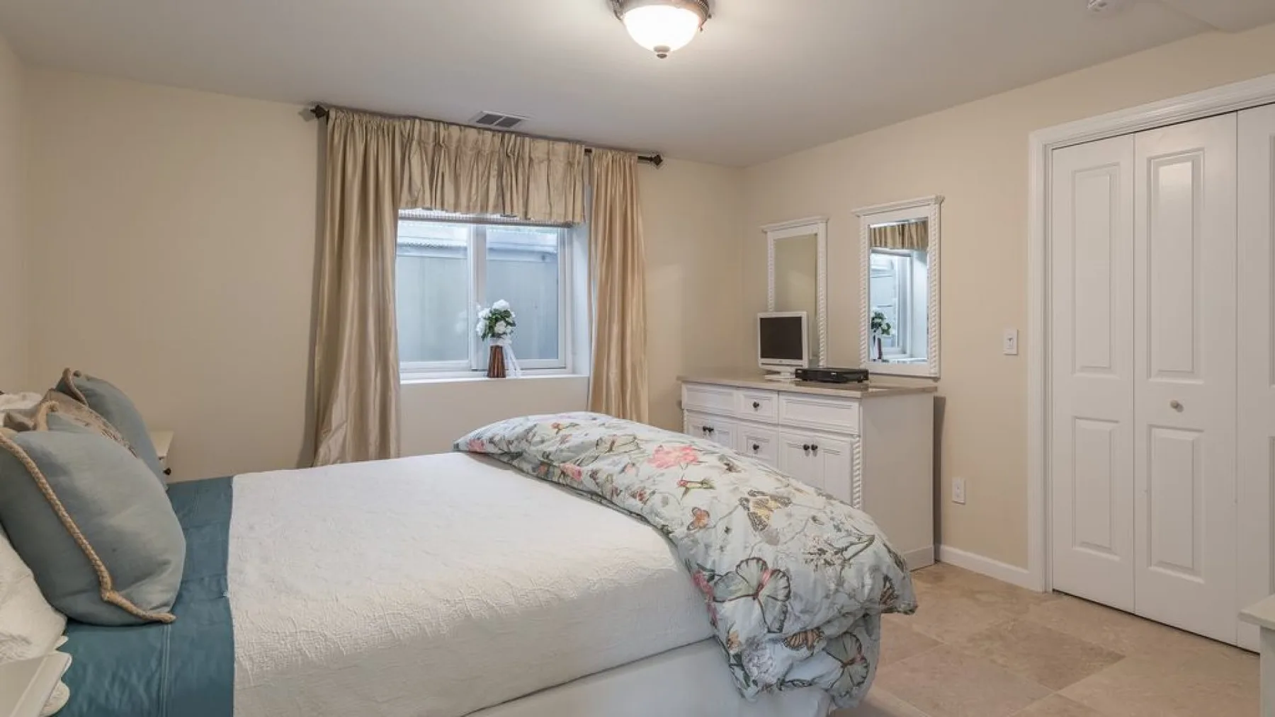 Un dormitorio con una cama, ropa de cama floral, una cómoda y una ventana con cortinas.