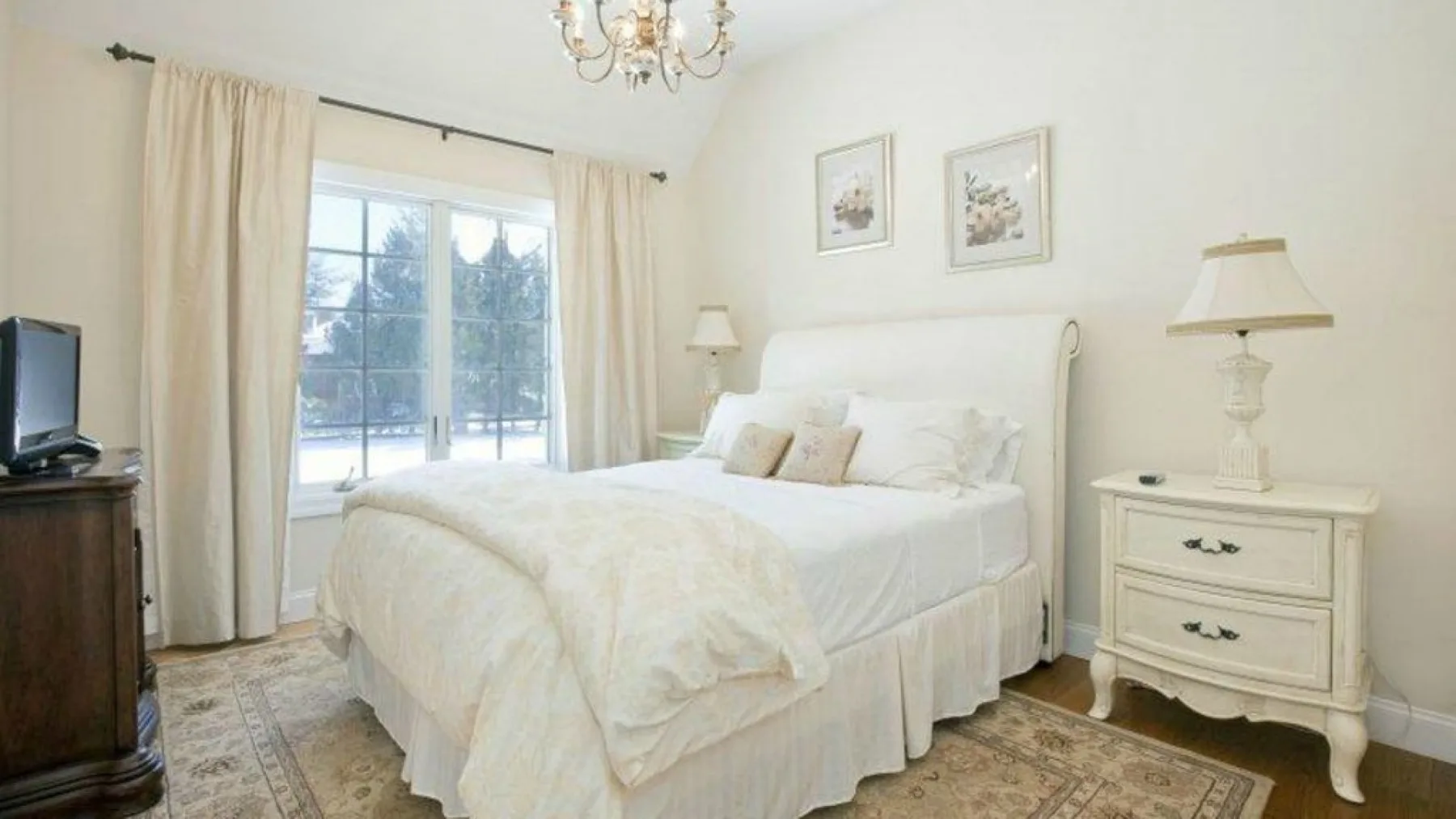 Un dormitorio luminoso con una cama junto a una ventana grande y cortinas color crema.