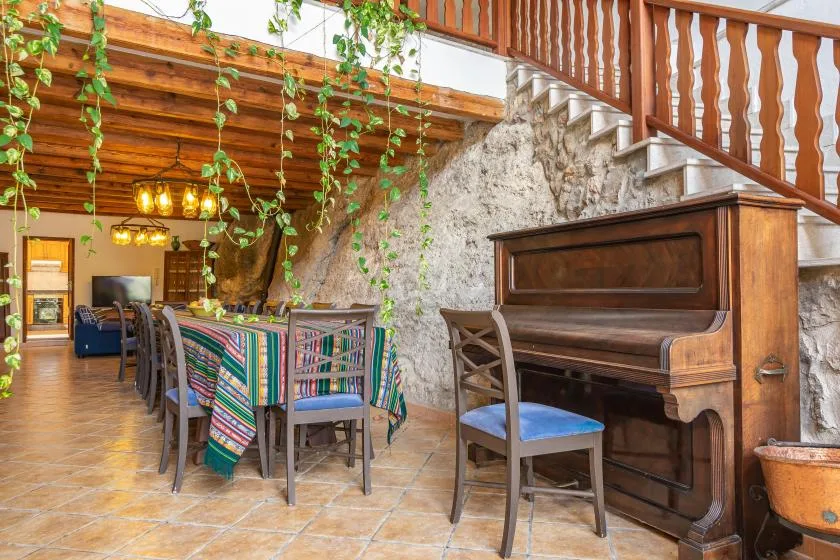 A rustic dining area with a long table and an upright piano beneath a wooden staircase.