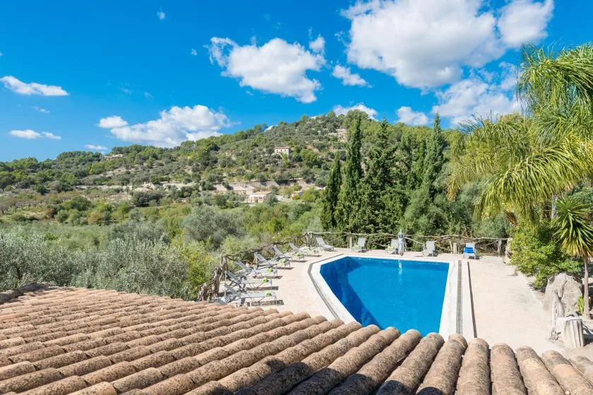 An outdoor swimming pool overlooks a green hillside beneath a blue, cloud-filled sky.