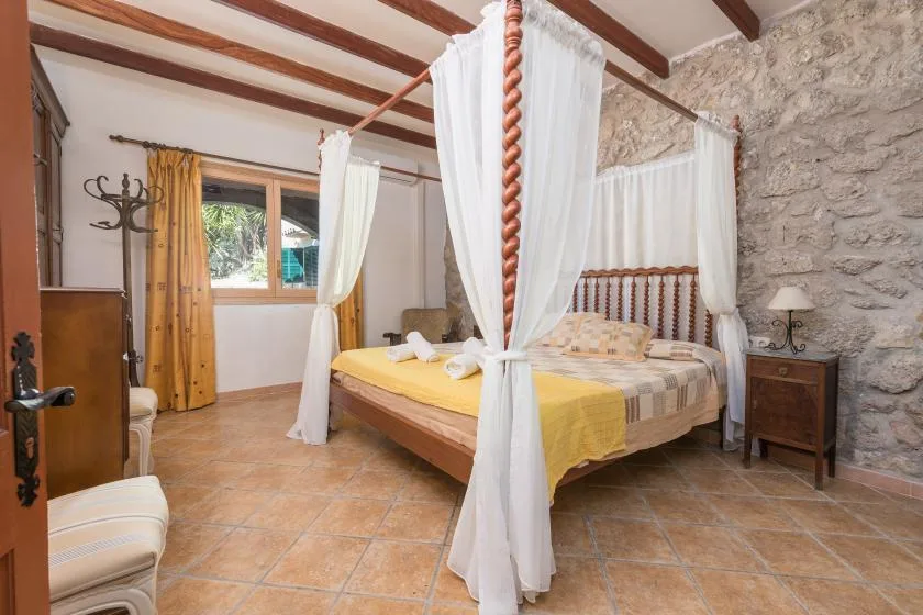 A bedroom with a wooden four-poster bed, white curtains, and a stone wall.
