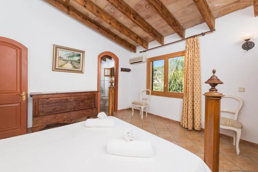 A bedroom with a bed, wooden ceiling beams, and a window overlooking greenery.