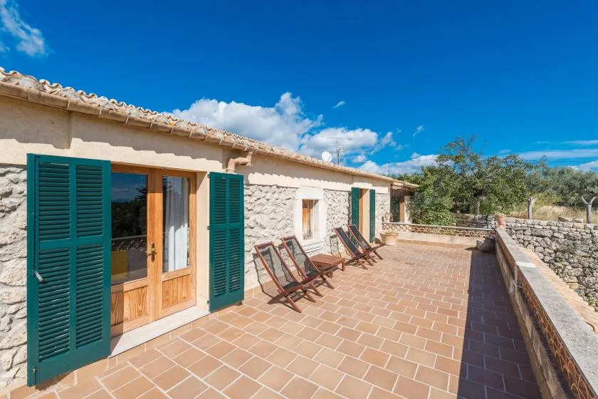 A sunny stone-house terrace with green shutters and outdoor seating.