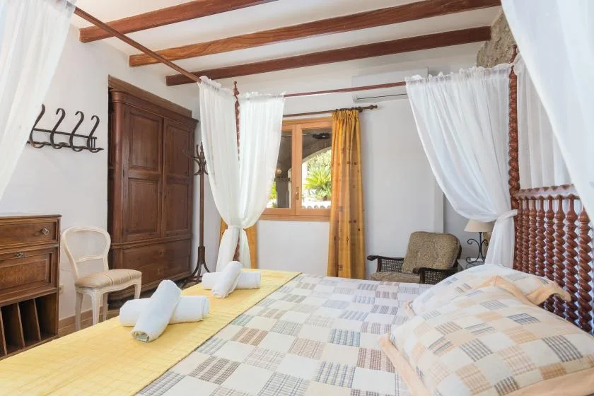 A bedroom with patterned bedding, wooden ceiling beams, and sheer curtains around the bed.