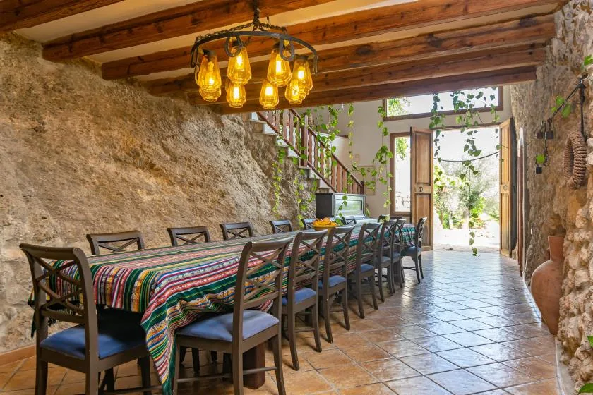 A long dining table with chairs sits in a rustic stone room beneath exposed wooden beams.