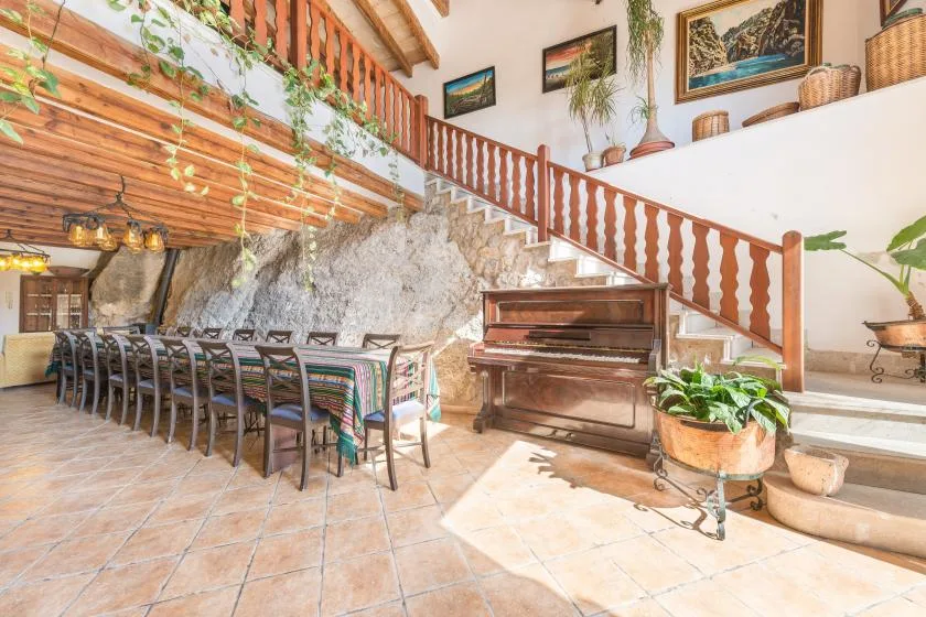 A dining area with a long table sits beside a piano beneath a staircase.