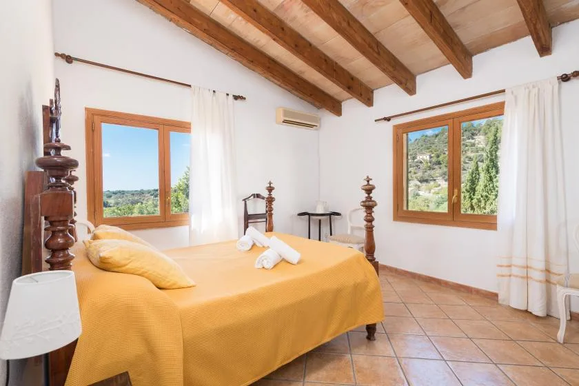 A bedroom with a yellow bedspread, exposed wooden ceiling beams, and two windows.