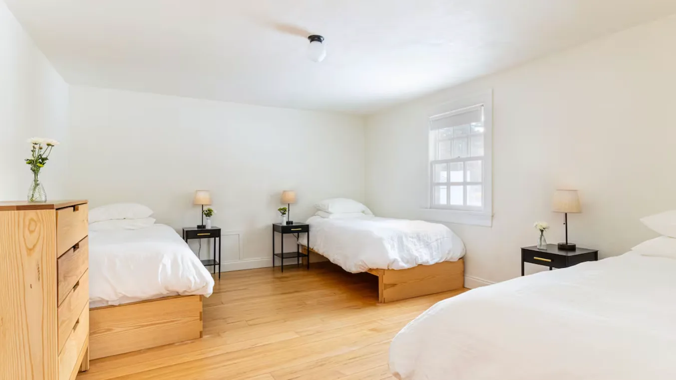 A bright bedroom with several beds, white bedding, and light wood floors.