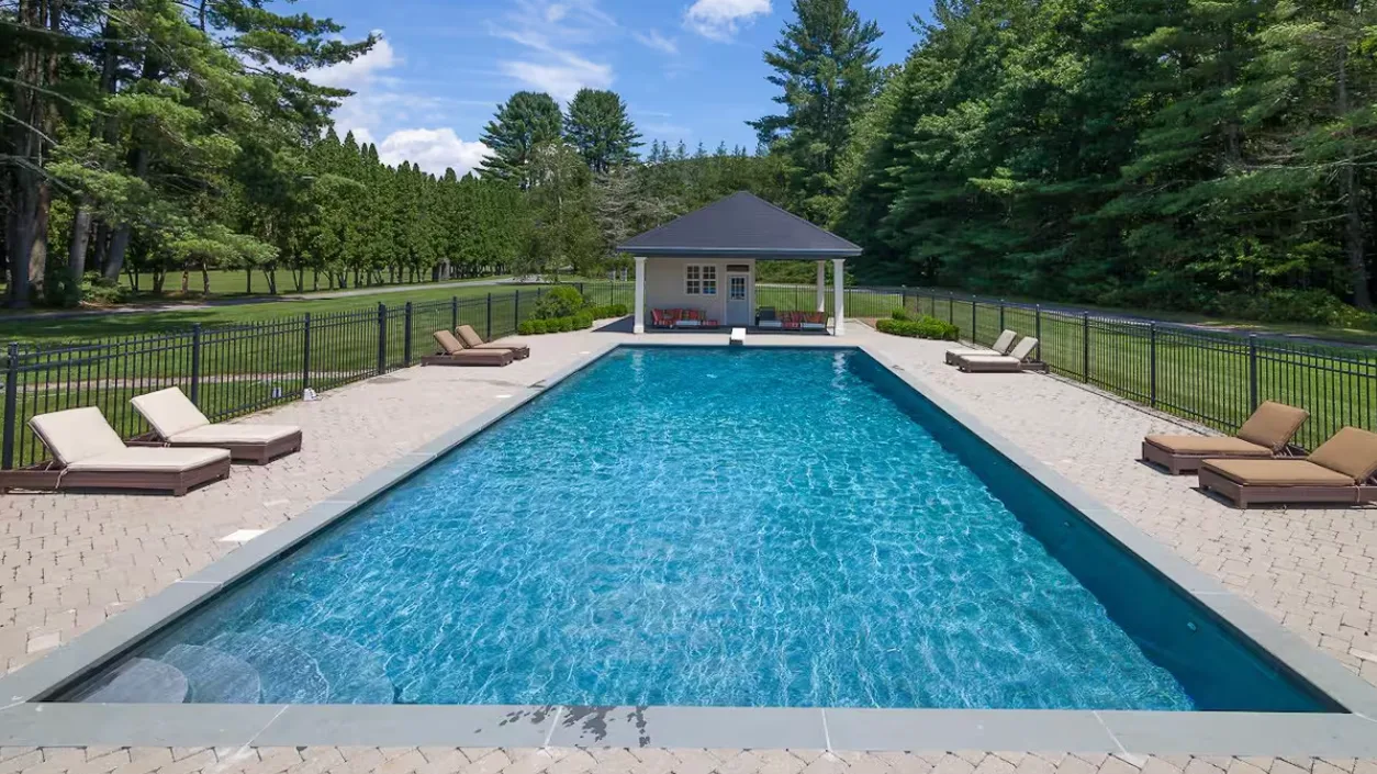 A swimming pool sits between paved decks, with trees surrounding the fenced area.