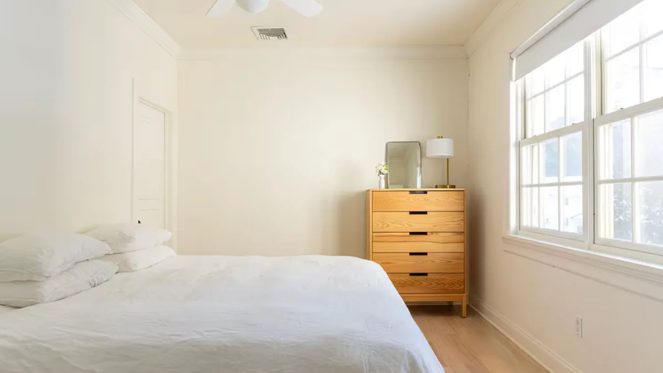 A bright bedroom with a bed, wooden dresser, and large windows.