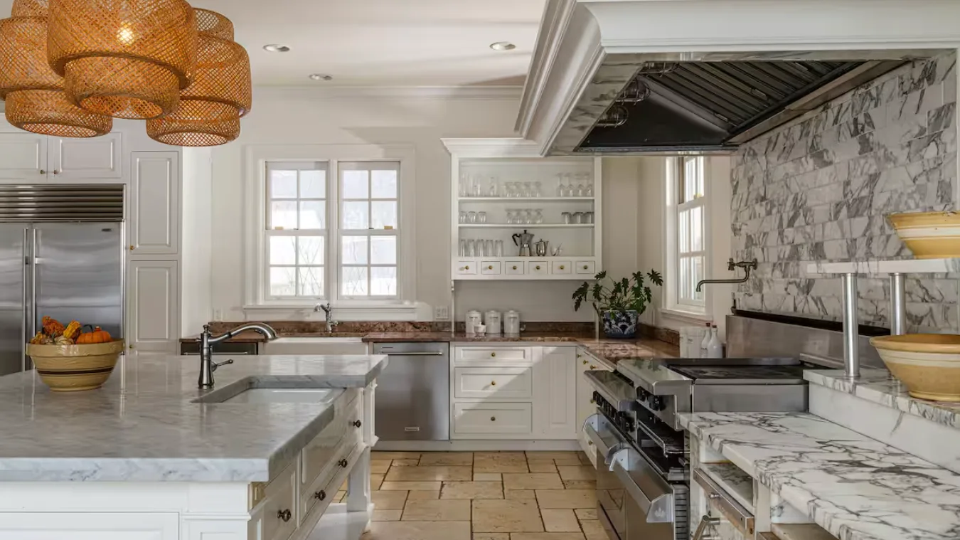 A spacious kitchen with marble countertops, white cabinetry, and a large range hood.