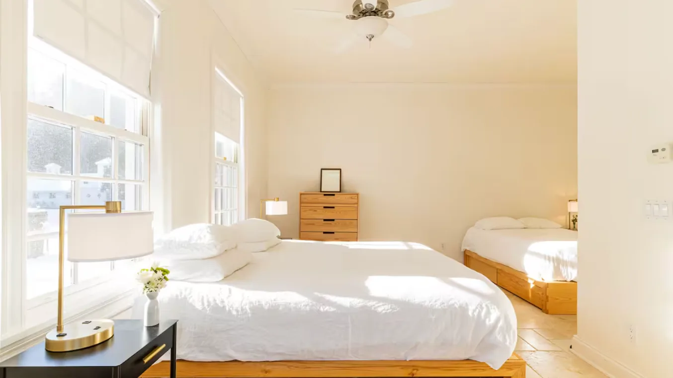 A bright bedroom with white bedding and large windows along one wall.
