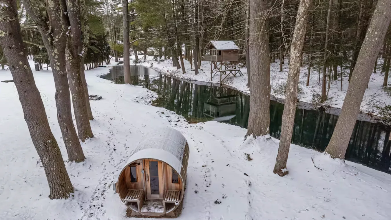 A snow-covered wooded area surrounds a small rounded cabin beside a stream.