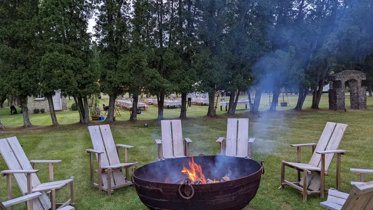 A grassy outdoor seating area surrounds a lit fire pit beneath tall trees.