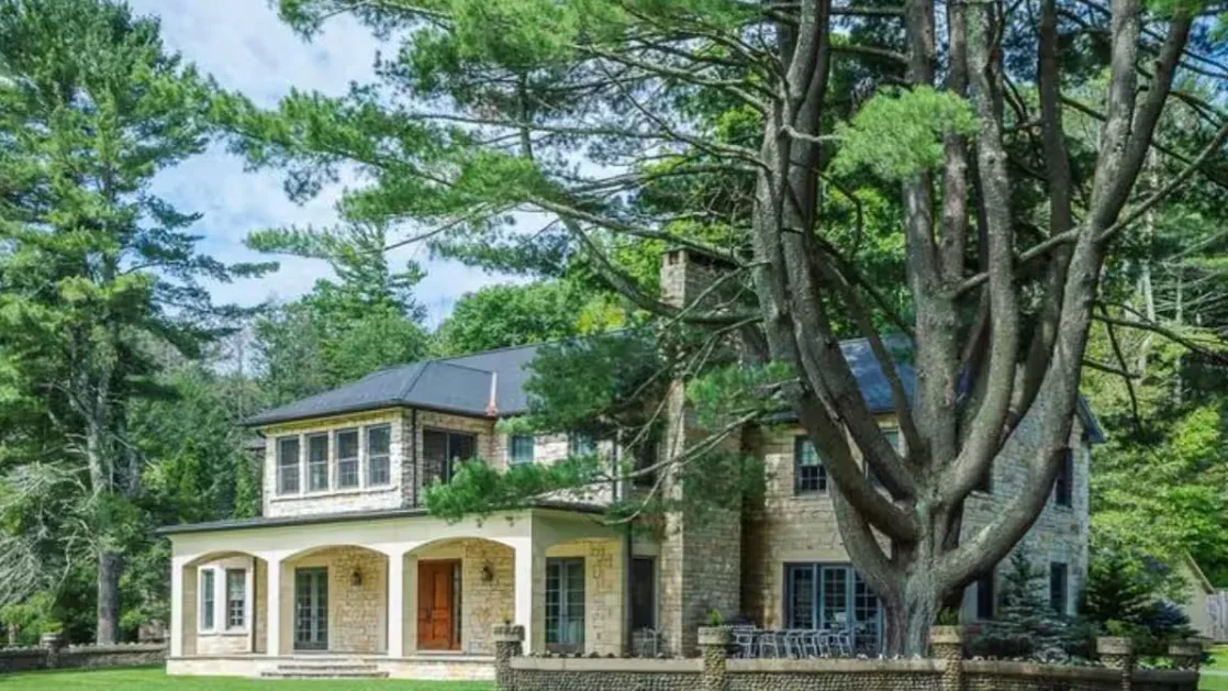 A large stone house stands among tall trees, with a covered entrance.