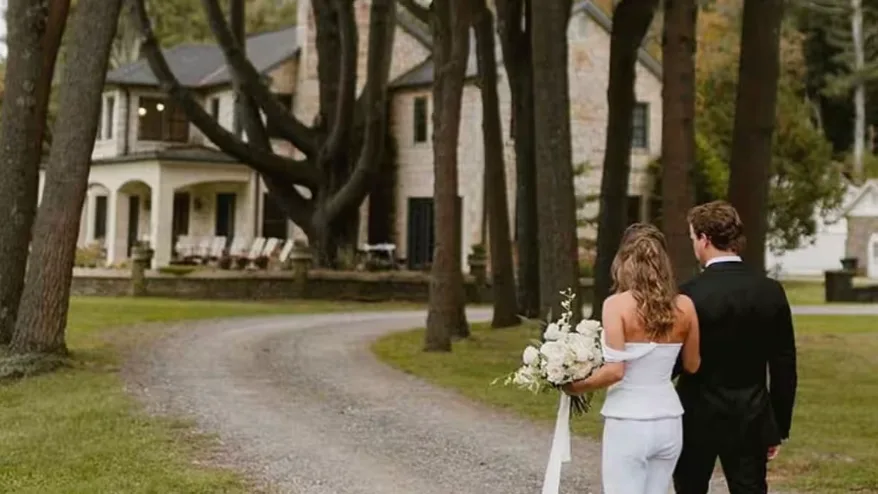 A couple walks along a gravel drive toward a large house, carrying a bouquet.