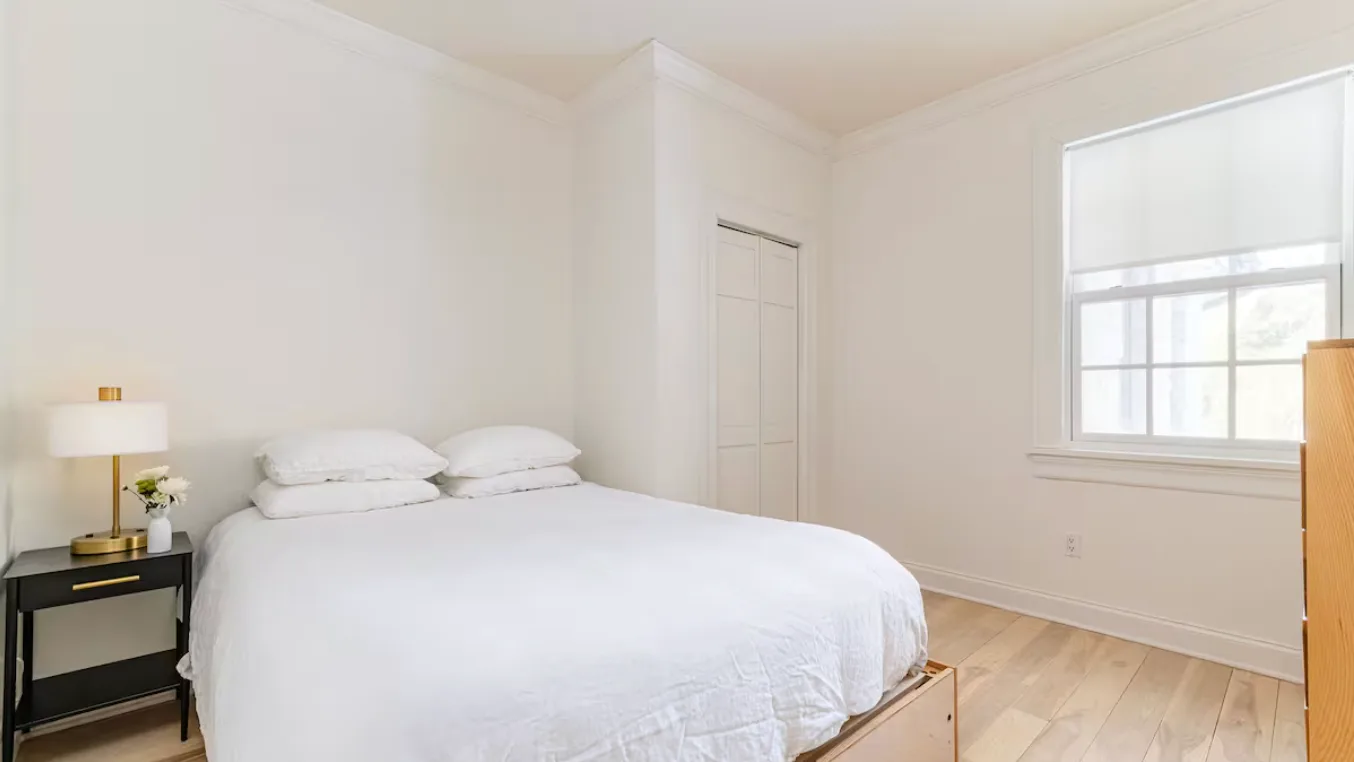 A bright bedroom with a white bed and a large window.