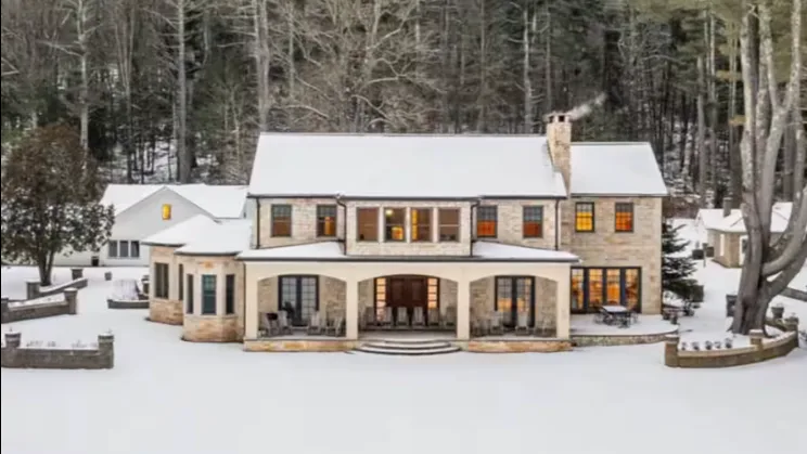A large house stands in a snowy clearing beside a wooded area.