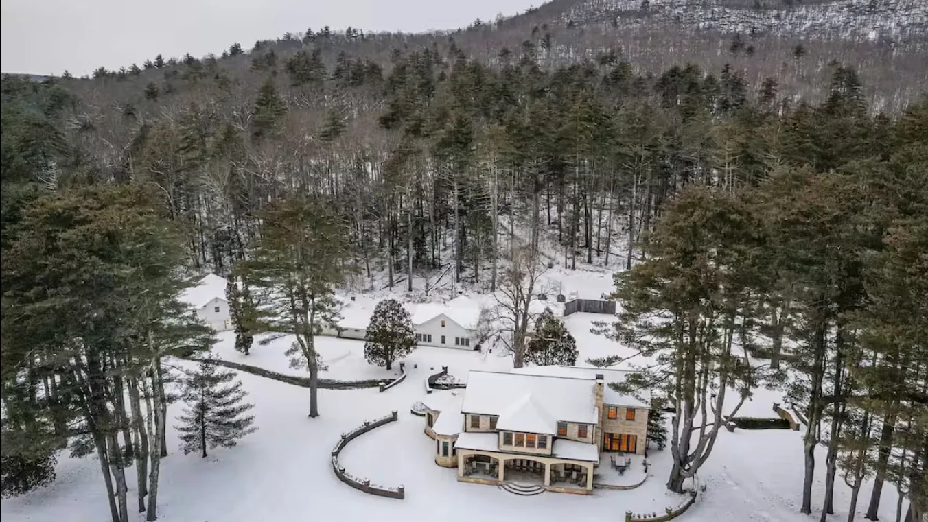 A snow-covered house sits among evergreen trees below a wooded hillside.