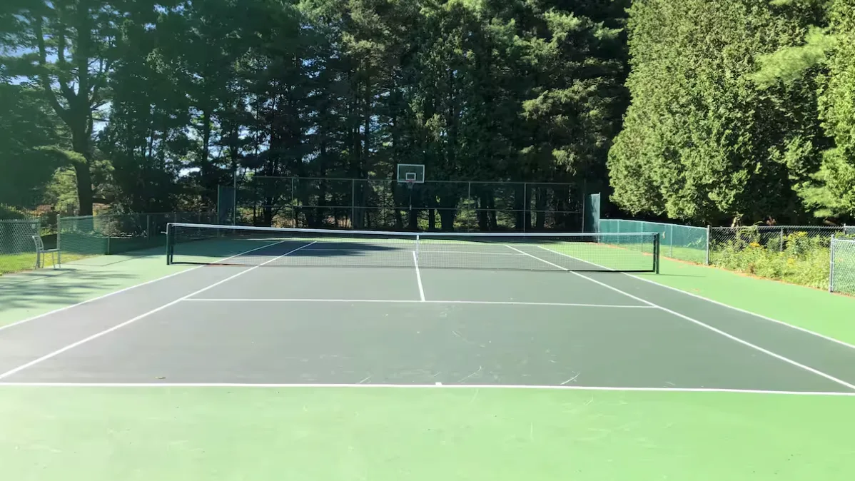 An outdoor tennis court with a basketball hoop behind the far end.