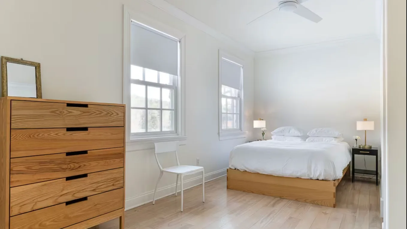 A bright bedroom with a bed, wooden dresser, and windows.