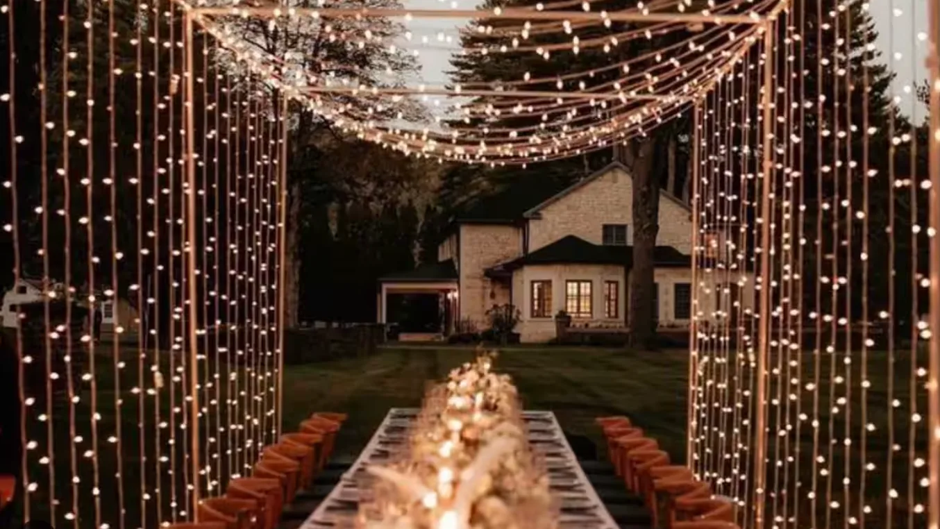 An outdoor dining area is lit by strands of lights, with a house in the background.