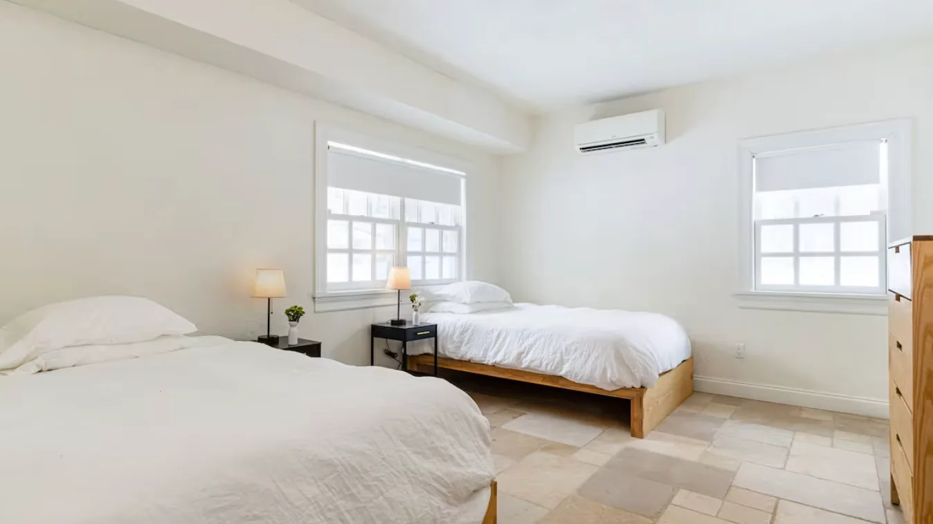 A bright bedroom with two beds, bedside tables, and windows.