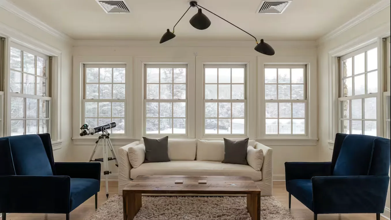 A bright living room has a sofa, blue seating, and a telescope by the windows.