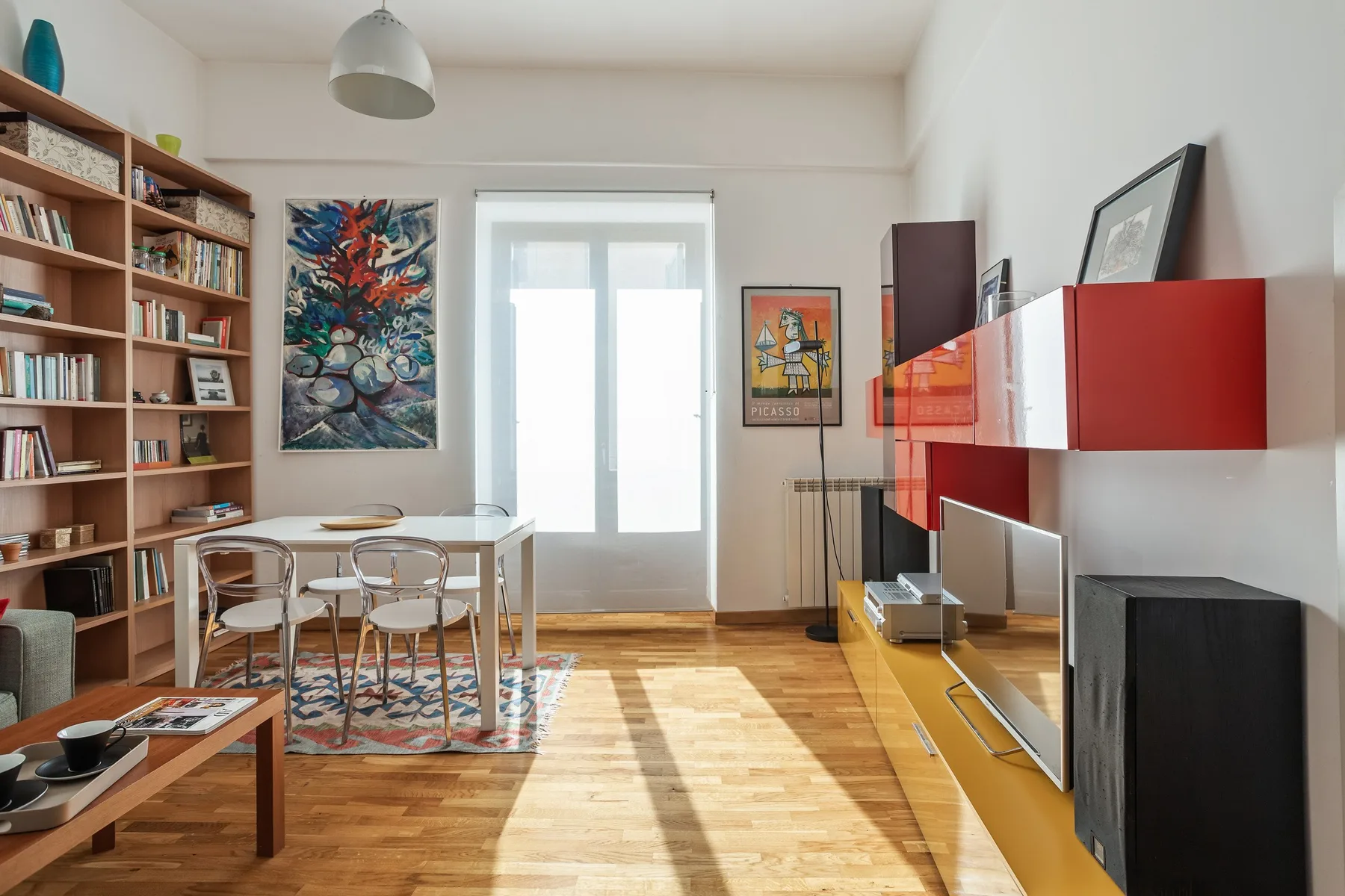 A bright living room with a dining table, bookshelves, and a wall-mounted television.