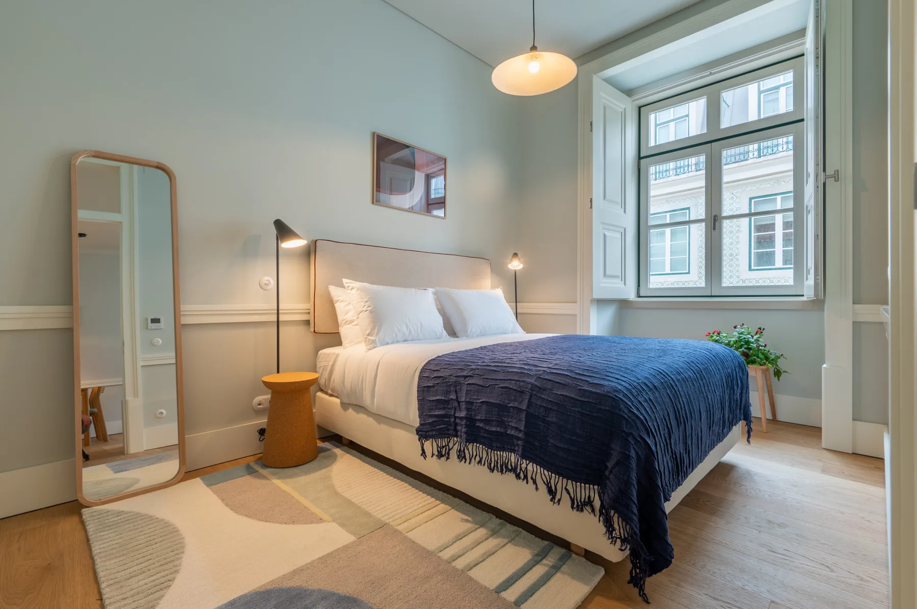 A bedroom with a bed dressed in white and a dark blue blanket beside a large window.