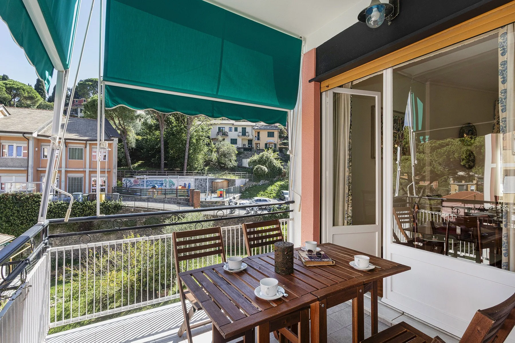 Un comedor en el balcón con mesa de madera y toldo verde, frente a edificios cercanos.