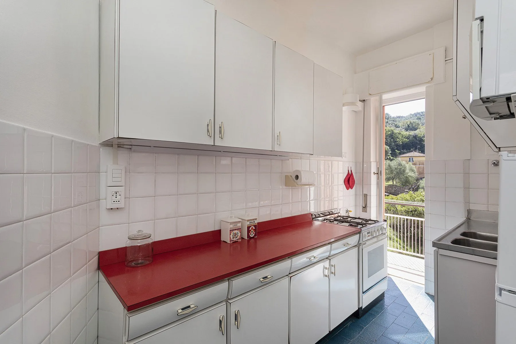 Una cocina luminosa con gabinetes blancos, una encimera roja y una puerta abierta al balcón.
