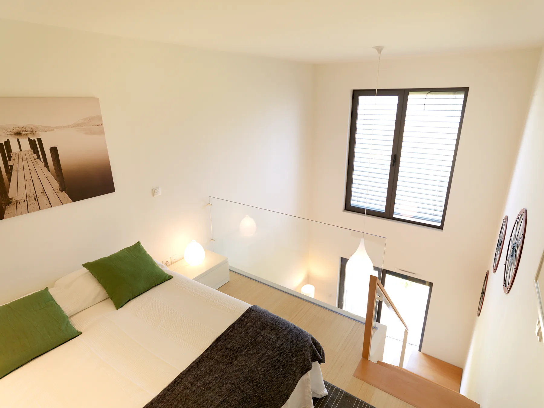 A bright loft bedroom has a bed beside a large window and open stairwell.
