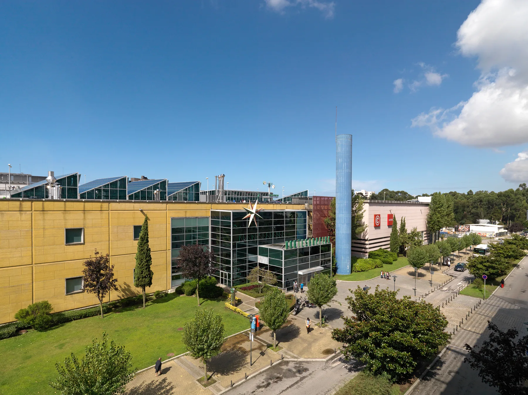 A modern glass-fronted building stands beside a landscaped lawn and tree-lined street.