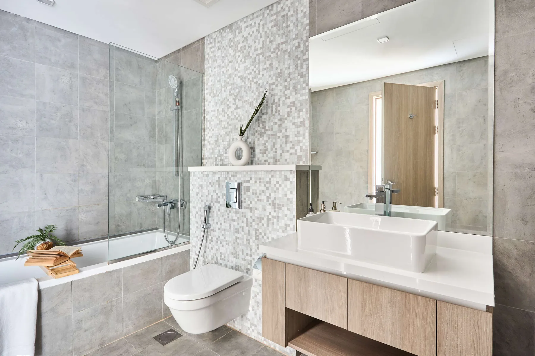 A bright tiled bathroom features a bathtub, wall-mounted toilet, and broad vanity.