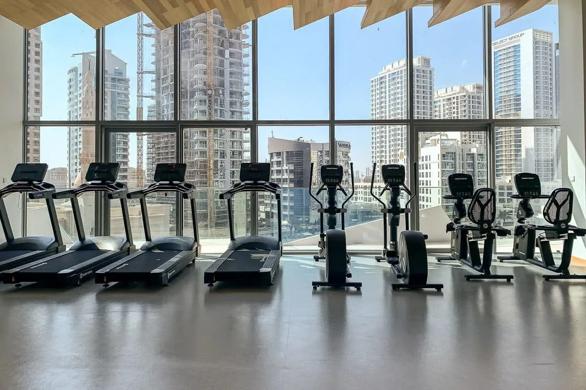 A bright fitness room with exercise machines in front of large windows.