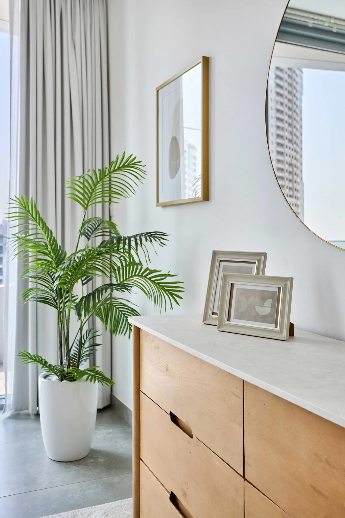 A bright room corner with a wooden dresser, framed pictures, and a potted palm.
