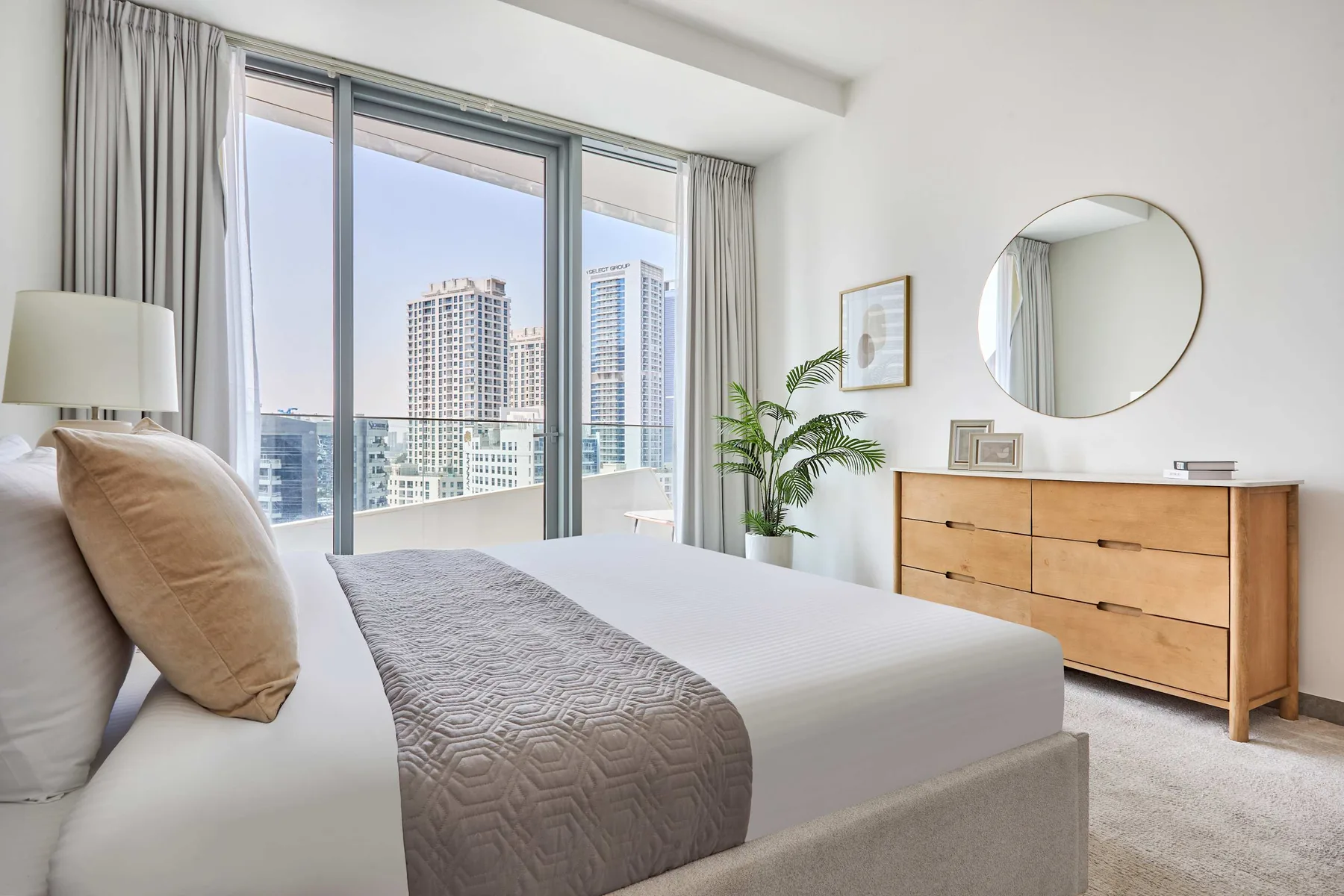 A bright bedroom with a neatly made bed beside large windows overlooking city buildings.