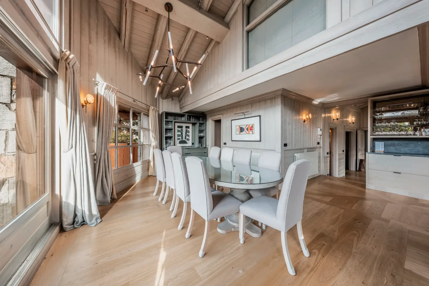 Spacious dining room with a long table, white chairs, large windows, wooden floors, and a vaulted ceiling with a modern chandelier.