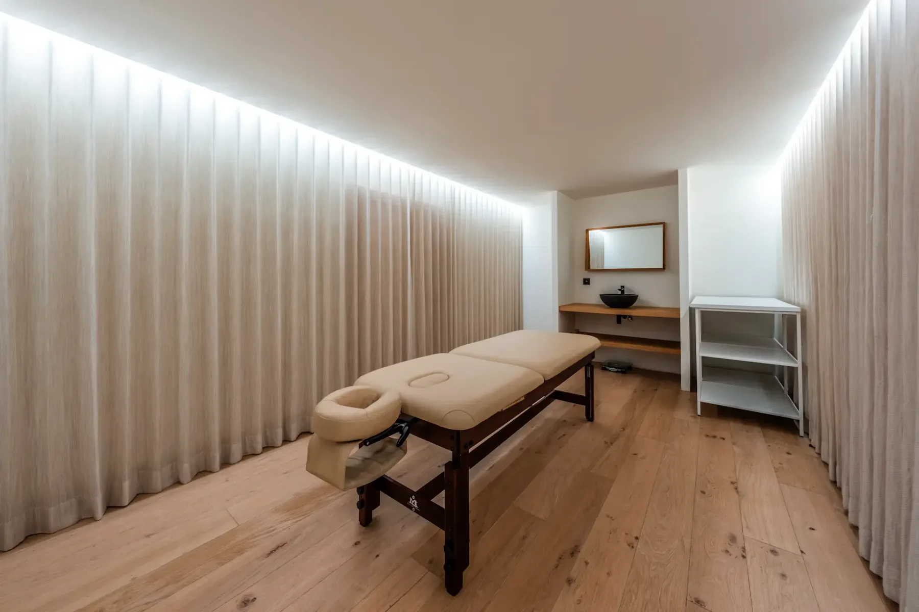 Minimalist massage room with a beige massage table, wooden floor, soft lighting, a mirror, and a small sink on a wooden shelf.
