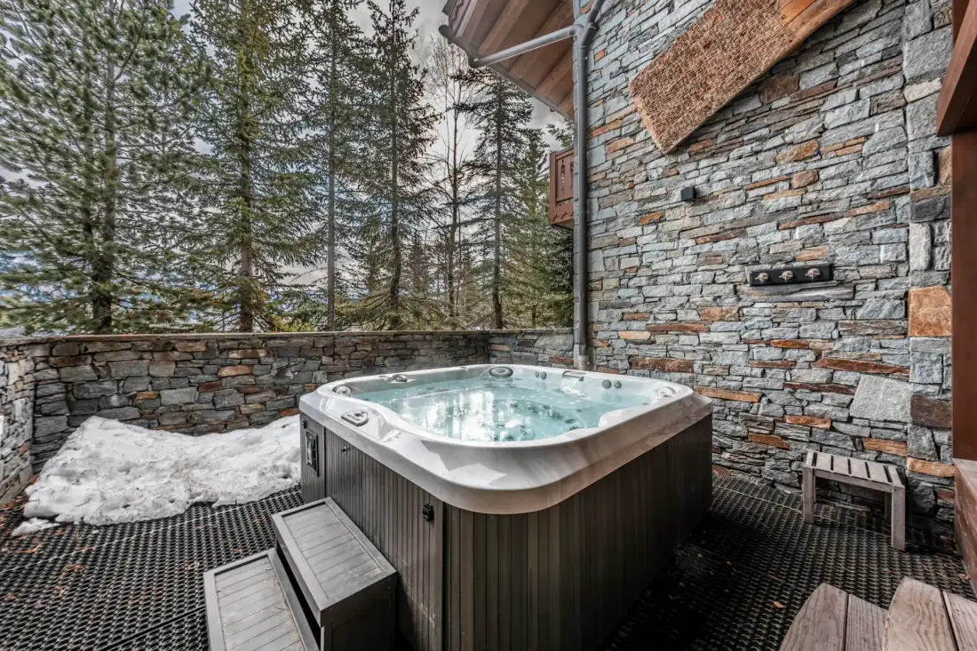 Outdoor hot tub on a stone patio surrounded by tall pine trees and snow, with a rustic stone wall and wooden elements.